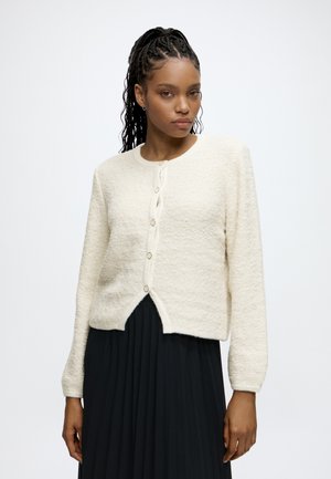Frau mit geflochtenem Haar trägt eine weiße, geknöpfte Strickjacke und einen schwarzen plissierten Rock, steht vor einem einfachen hellen Hintergrund.
