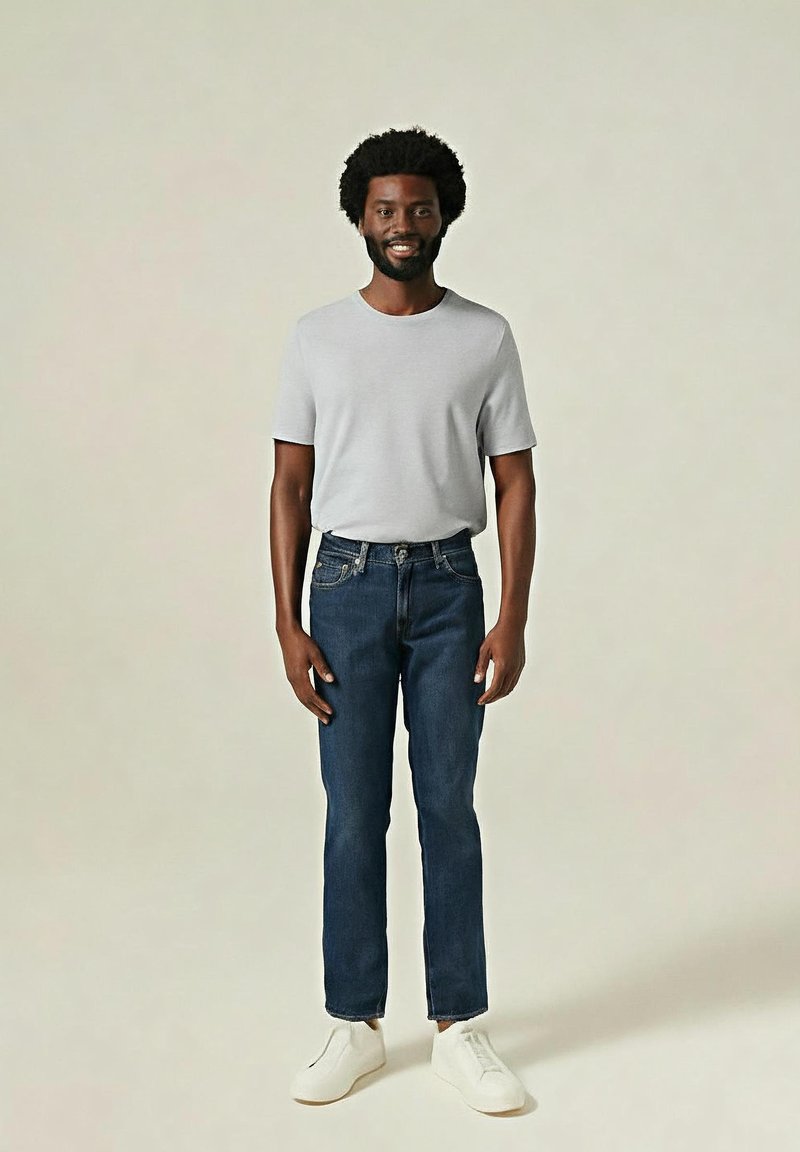 Lächelnder Mann mit Afrofrisur, der ein hellgraues T-Shirt, dunkelblaue Jeans und weiße Sneakers trägt, vor einem einfarbigen Hintergrund stehend.