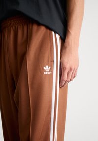 Brūnas sporta bikses ar elastīgu jostasvārstu, baltām sānu strīpām un izšūtu baltu Adidas logo kreisajā augšstilbā.
