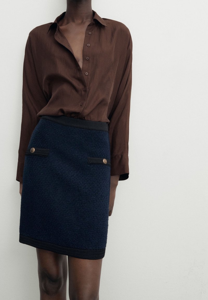 Femme portant une chemise marron foncé boutonnée et une jupe bleu marine texturée avec une bordure noire et des détails de boutons dorés sur les poches avant.