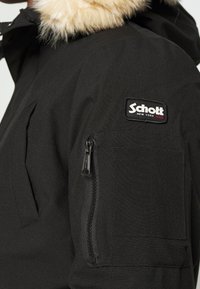 Veste d'hiver noire avec une capuche bordée de fourrure. Elle possède une poche zippée sur la manche gauche et un logo Schott New York sur la poitrine.