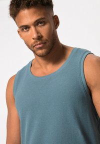 Top in cotone teal con texture a coste, ampie aperture per le braccia e collo girocollo classico, esposto su uno sfondo chiaro.
