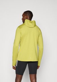 Langärmliges Kapuzenshirt in leuchtendem Limettengrün. Weiche, glatte Textur mit tailliertem Design. Verfügt über ein dezentes Logo und ist mit schwarzen Shorts kombiniert.
