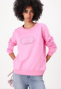 Sweatshirt rose avec un col rond, des manches froncées et un design de logo avec des accents métalliques. Associé à un jean bleu clair.