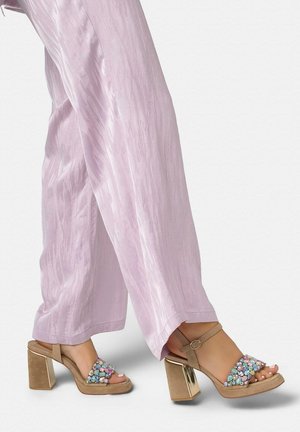 Pies con sandalias de tacón beige con gemas multicolores y tiras en el tobillo, combinadas con pantalones de pierna ancha color rosa claro.