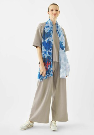 Vrouw met beige losse broek en T-shirt, witte sneakers en een lange blauwe sjaal met hartjespatroon, staand tegen een witte achtergrond.