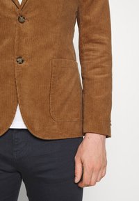 Hnědý kordový blazer s klopovým límcem, dvěma knoflíky, jednou náprsní kapsou a texturovanou látkou. Nositý přes bílé triko a tmavé kalhoty.