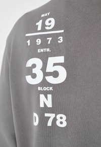 Grå sweatshirt med stort hvidt trykt tekst: "MAY 19 1973 ENTR. 35 BLOCK N D 78." Teksten er fed med en tekstureret baggrund.