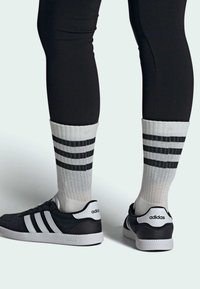 Zapatillas deportivas negras con tres rayas blancas, combinadas con calcetines grises que tienen tres rayas horizontales negras y un puño acanalado.