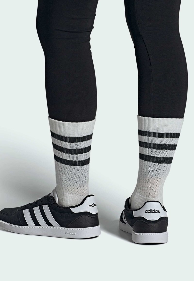 Zapatillas deportivas negras con tres rayas blancas, combinadas con calcetines grises que tienen tres rayas horizontales negras y un puño acanalado.