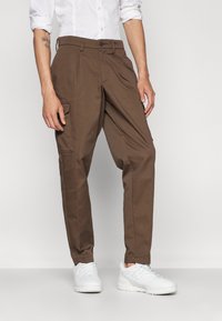 Brown cargo pants with a tailored fit, made of sturdy fabric. Features side pockets, a zip fly, and a button closure. Pair with white sneakers.