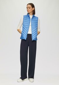 Gilet matelassé bleu clair avec fermeture éclair et coutures horizontales, porté sur un haut à manches longues blanc et associé à un pantalon large sombre.
