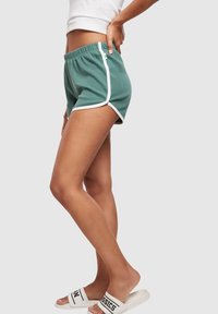 Person wearing green athletic shorts with white trim and white slides, standing against a plain light gray background.