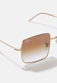 Obdélníkové sluneční brýle s gradientními hnědými čočkami, zlatým kovovým rámem a průhlednými konci nožiček. Logo Ray-Ban na čočce. Lehký design.