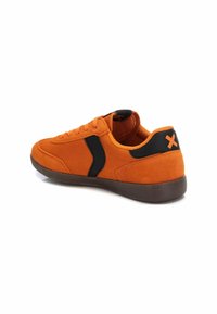 Zapatillas de ante naranja con acentos negros, cordones planos y una suela de goma marrón texturizada. Presenta un logo en el talón y en el panel lateral.