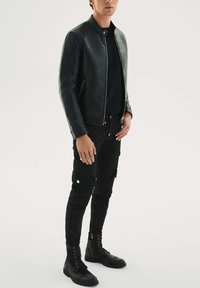 Črna usnjena jakna z visokim ovratnikom in zadrgo, kombinirana s črnimi cargo hlačami in čevlji s vezalkami v vojaškem slogu, ki prikazuje eleganten dizajn.