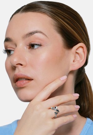 Frau mit leichtem Make-up und zurückgebundenen Haaren, die ihr Kinn berührt, trägt einen silbernen Ring, der wie ein Blatt geformt ist, am Finger.