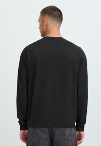Mann med kort hår og en øredobb iført en enkel svart langermet skjorte og mørke jeans, vendt bort fra en lys bakgrunn.