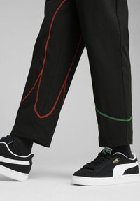 Pantalones negros con costuras en contraste rojas y verdes, combinados con unas zapatillas negras y blancas de ante con una superficie texturizada.