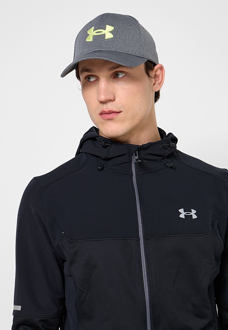 Jeune homme regardant vers la droite, portant une casquette grise avec un logo néon et un sweat à capuche zippé noir avec un logo de marque réfléchissant sur la poitrine.