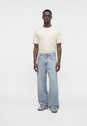 Mann steht vor schlichtem Hintergrund, trägt ein blassgelbes T-Shirt, hellblaue weite Jeans und silberne Sneaker.