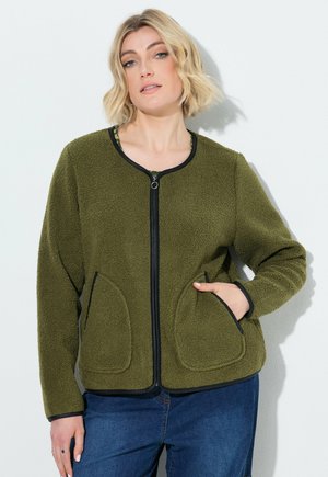 Mujer rubia con chaqueta texturizada con cremallera color verde oliva, con ribetes negros y bolsillos delanteros, combinada con vaqueros azules, sobre fondo blanco.