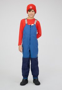 Wasserfester Overall in Blau und Marineblau mit verstellbaren grauen Trägern. Darunter ein langärmliges rotes Shirt, schwarze Gummistiefel und eine rote Strickmütze.