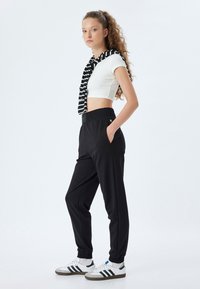 Schwarze Jogginghose mit elastischem Bund und Bündchen, kombiniert mit einem weißen gerippten Cropped-Top und einem schwarz-weißen gestreiften Schal, der um den Hals gebunden ist.