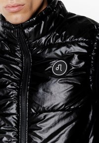 Czarna pikowana kurtka puchowa z wysokim kołnierzem, błyszcząca faktura, pełny zamek błyskawiczny oraz okrągła naszywka z logo na lewej części klatki piersiowej.