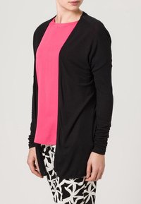Czarny kardigan z długim rękawem, otwarty z przodu, nałożony na jasnoróżową bluzkę. Połączony z czarno-białymi spodniami w wzory o gładkiej fakturze.