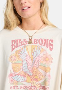 Cream-colored oversized t-shirt featuring a retro graphic with an eagle, flowers, and text in pink and blue hues. Gold chain necklace with pendant.