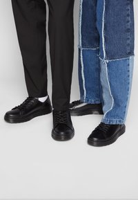 Baskets noires avec une tige en cuir lisse, bout rond et semelles texturées, associées à un pantalon en denim noir et patchwork.