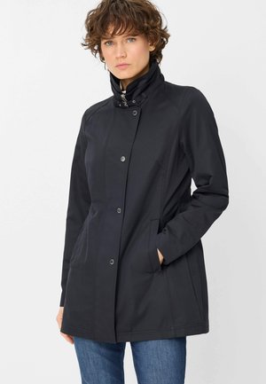 Junge Person mit kurzen lockigen Haaren, die eine schwarze Jacke mit hohem Kragen und Druckknöpfen trägt und die Hände in den Taschen hat, vor einem weißen Hintergrund stehend.