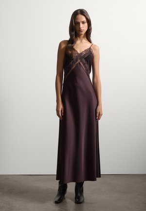 Donna con i capelli lunghi e scuri che indossa un vestito sottoveste in raso viola scuro con dettagli in pizzo e stivaletti neri, in piedi davanti a uno sfondo chiaro e uniforme.