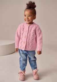 Cardigan rosa lavorato a maglia con motivi a treccia, bottoni in legno e polsini a costine; abbinato a jeans di denim blu e stivaletti rosa chiaro.