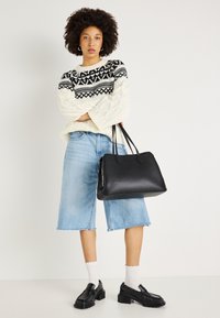 Mustmust kott kahe sangaga, sile tekstuur, ristküliku kuju. Seljas kreemjas nööridega kampsun ja helesinised teksast lühikesed püksid.