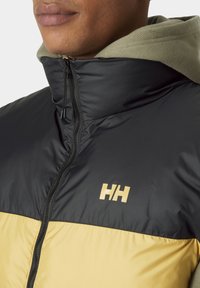 Polsterjacke mit einem schwarzen oberen Bereich und einem gelben unteren Teil, mit einem hohen Kragen, Reißverschluss und einem goldenen Logo auf der Brust.