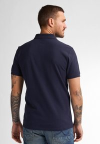 Námořnická polo tričko s krátkými rukávy, límečkem a bočními rozparky. Vyznačuje se hladkou texturou a přiléhavým střihem. Viditelné tetování na paži.