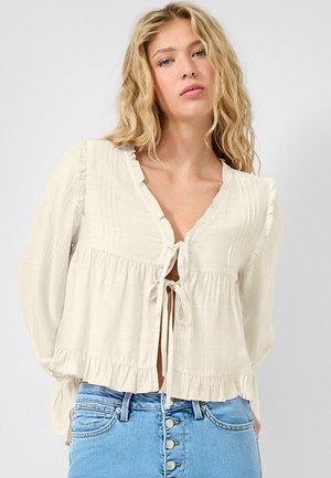 Femme aux cheveux blonds ondulés portant un chemisier crème à manches longues avec des liens à l'avant et un jean bleu taille haute à boutons, sur un fond uni.