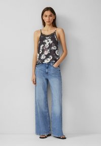 Camisola nera floreale con pizzo, abbinata a jeans a gamba larga blu chiaro. La modella è in piedi contro uno sfondo neutro, indossando sandali neri.