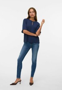 Donna in piedi con le braccia incrociate, che indossa una blusa blu navy, jeans attillati blu e mule con tacco a punta marroni su uno sfondo bianco.