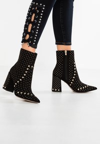 Musta mokkapohjaiset nilkkurit teräväkärkiset, koristeltu kultaisilla niiteillä, chunky-korko ja sivu vetoketju kiinnitystä varten.