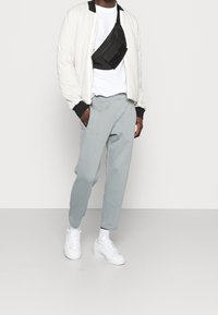 Vit bomberjacka, svart midjeväska, vit T-shirt, ljusgrå joggers och vita sneakers. Slät tyg med ribbade detaljer vid ärmarna.