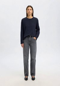 Nainen seisoo kasvot kohti kameraa, päällään meri- sininen pitkähiha- paita, harmaat korkeavyötäröiset farkut mustalla vyöllä ja mustat kärkitossut.