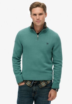 Teal zip-up sweater with ribbed collar and cuffs, layered over a black checkered shirt. Worn with dark jeans and a wristwatch.