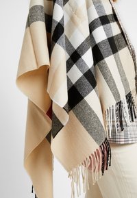 Châle à franges à carreaux beige, noir et blanc drapé sur une personne portant un pantalon crème et une veste à carreaux.