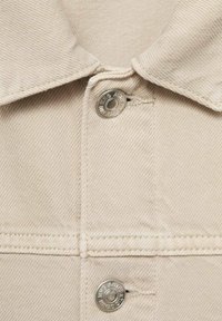 Giacca di jeans beige con colletto button-up e bottoni a pressione argentati. Il tessuto ha una finitura testurizzata con cuciture visibili.