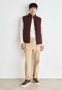 Uomo in piedi contro un muro bianco, indossando una camicia bianca con maniche lunghe, un gilet trapuntato bordeaux, pantaloni beige arrotolati e scarpe da ginnastica marroni.