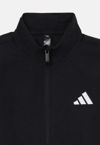 Giacca sportiva nera con colletto alto, zip completa e logo bianco con tre strisce sul lato sinistro. Texture liscia.