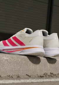 Hvide løbesko med pink striber og orange detaljer, der hviler på en betonkant i et udendørs miljø.
